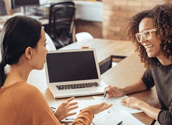 Two women collaborating at desk with laptop and documents in office setting