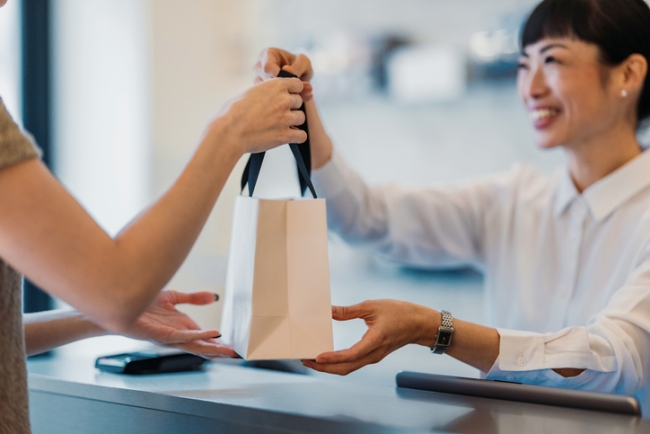  Smiling sales clerk handing a small white shopping bag to a customer over a counter.